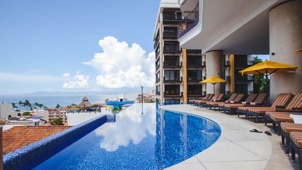 Una piscina con sillones de descanso y sombrillas, vista al océano y un edificio con balcones.