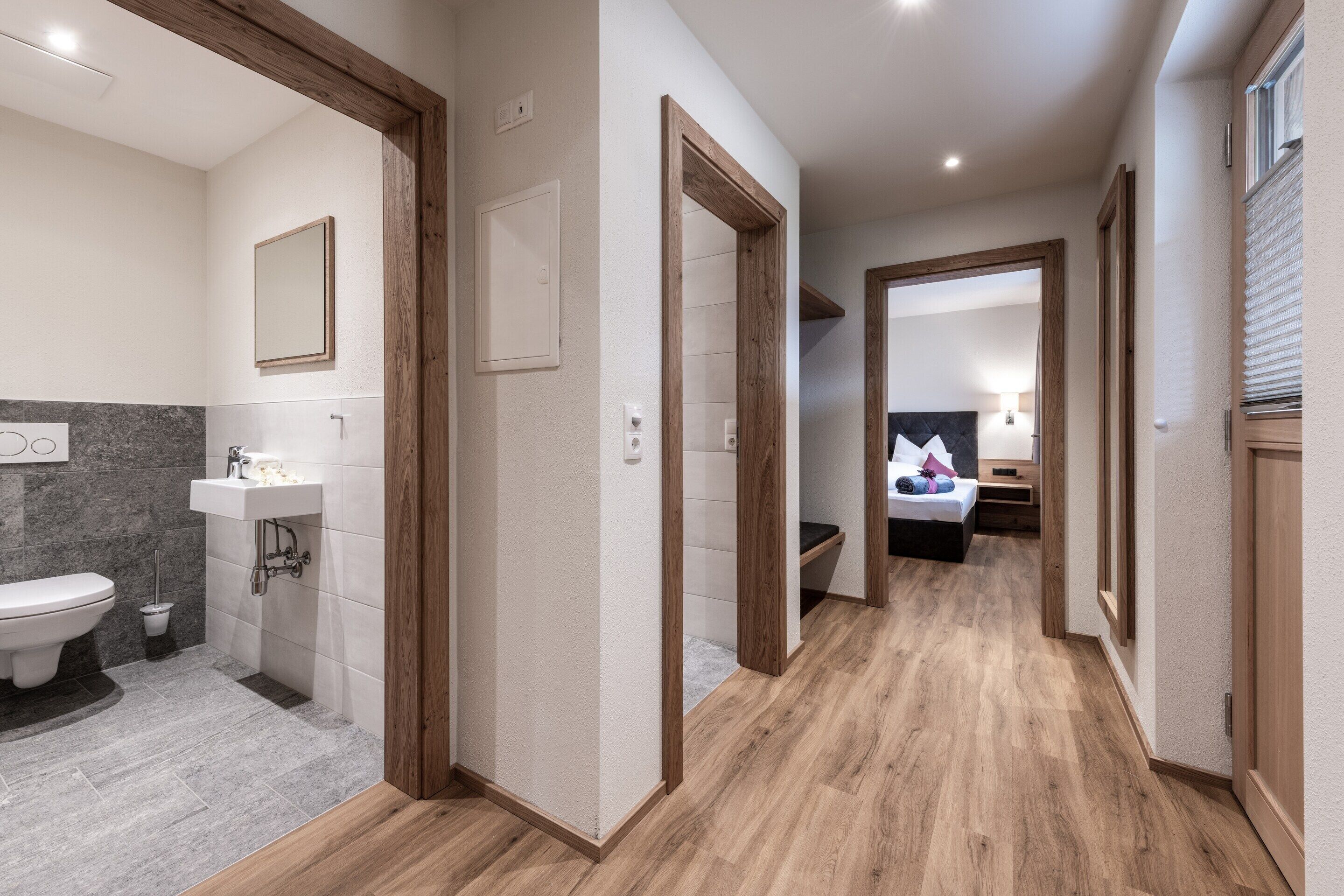 A modern hotel room with a wooden floor, a bathroom with a sink and toilet, and a bed with a headboard.