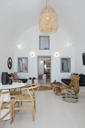 A modern living room with a round table, wicker chairs, a sofa, and a hanging light fixture. - You And Me Suites (Santorini)