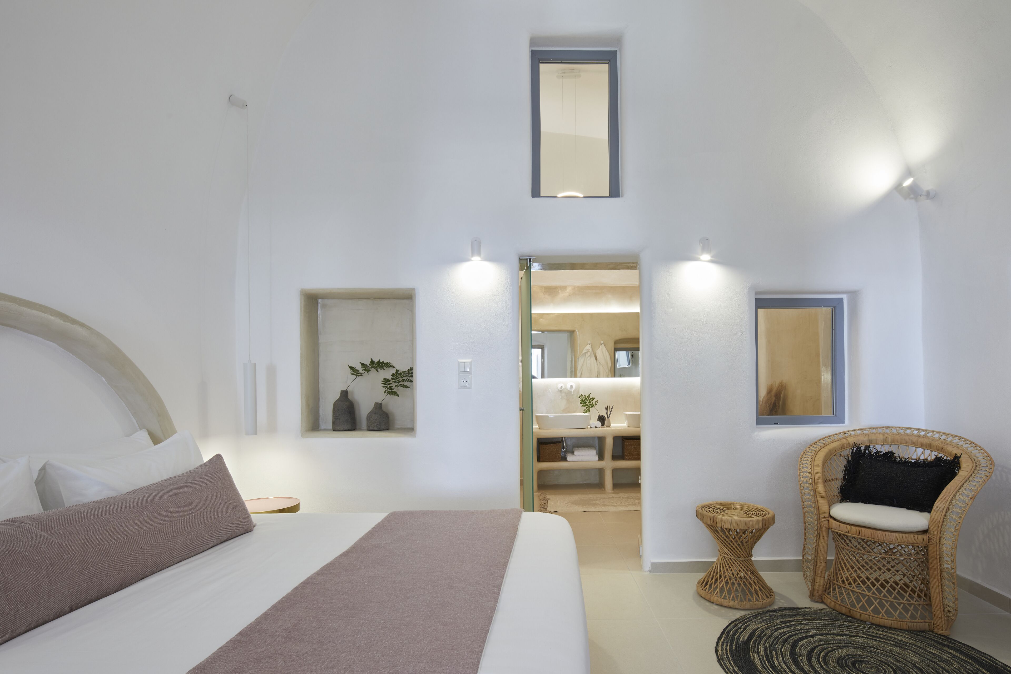 A modern bedroom with a large bed, a small side table, a wicker chair, and a bathroom visible through an open door.