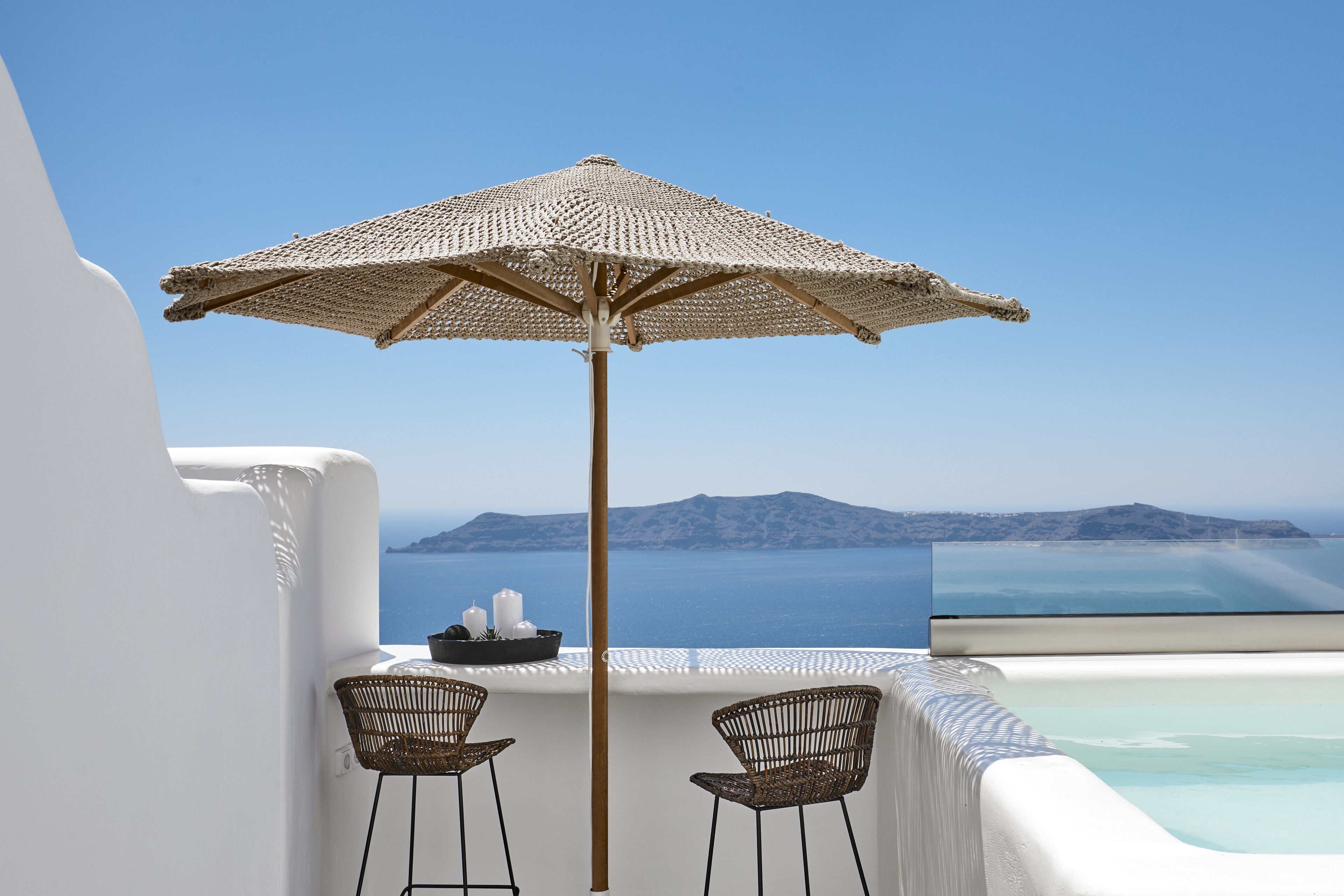 A balcony with a woven umbrella, two wicker chairs, a small table with candles, and a view of the sea and islands.