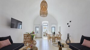 A modern living room with a sofa, a coffee table, a wicker chair, and a dining area with a table and chairs. - You And Me Suites (Santorini)