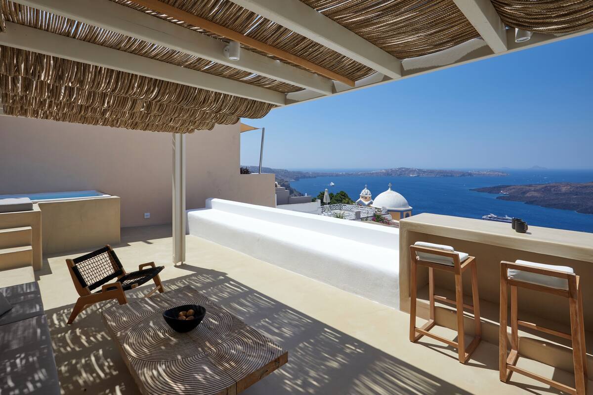 A terrace with a view of the sea, a white bench, a wooden chair, and a table with a bowl.