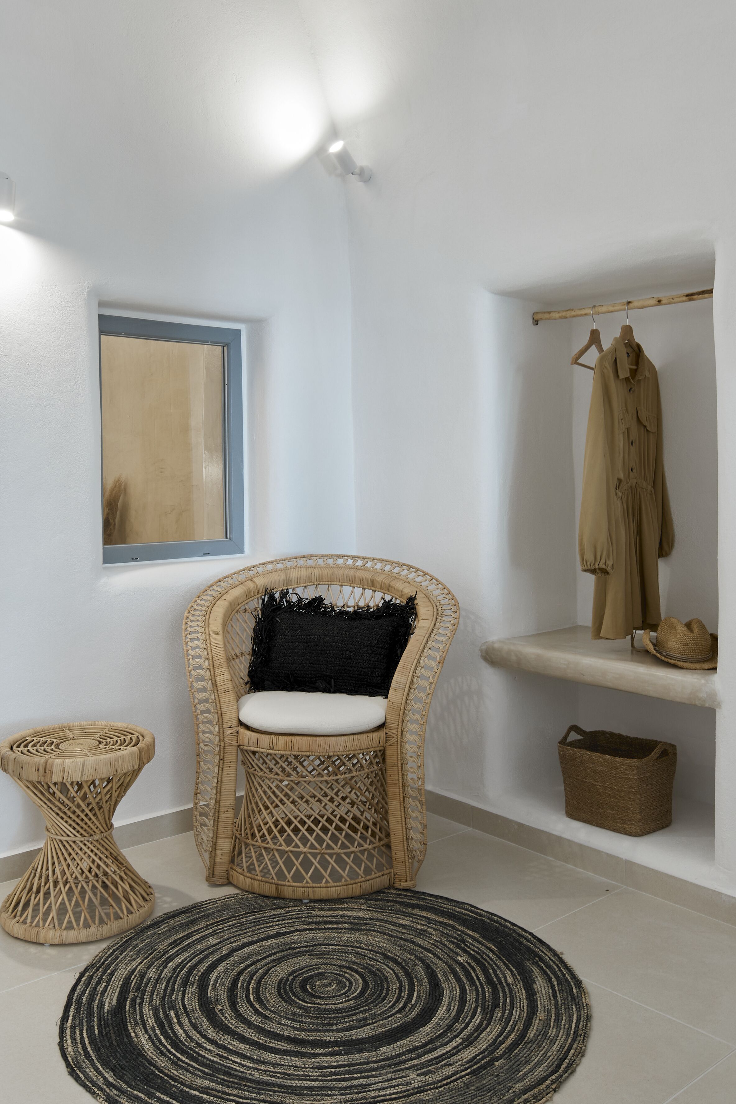 A room with a wicker chair, a round black and white rug, a small side table, a coat rack with a coat, and a framed picture on the wall.