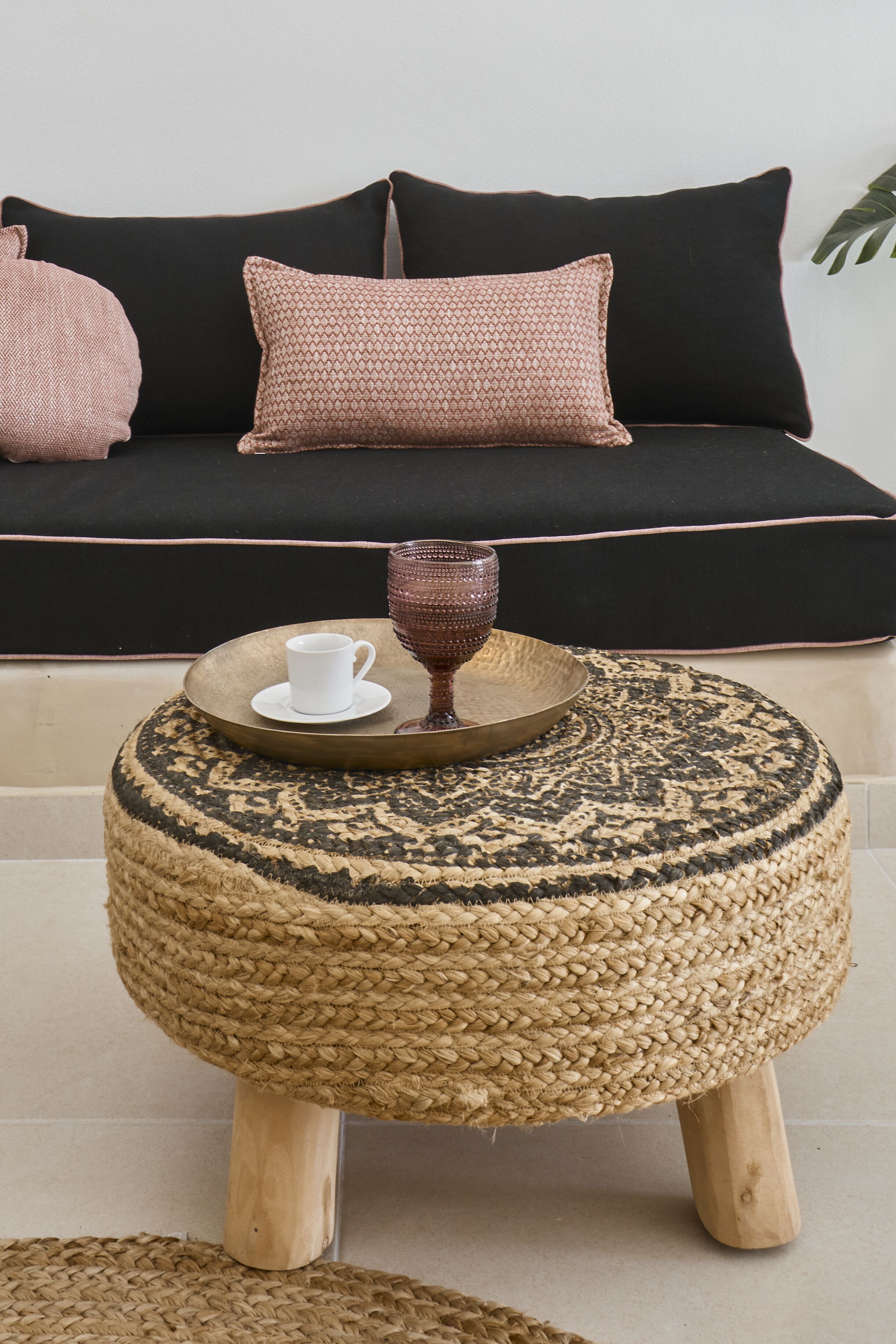 A round woven ottoman with a tray and cup on top, placed in front of a black sofa with pink cushions.