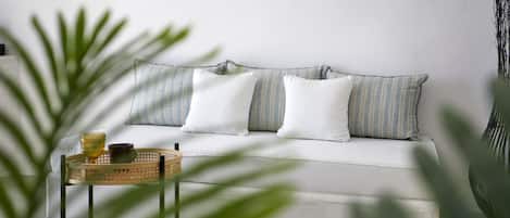 A white sofa with striped pillows, a small wooden side table with a glass and a cup, and a large green plant in the foreground.