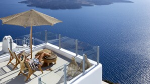 A balcony with a view of the sea, wicker furniture, and a glass railing. - You And Me Suites (Santorini)