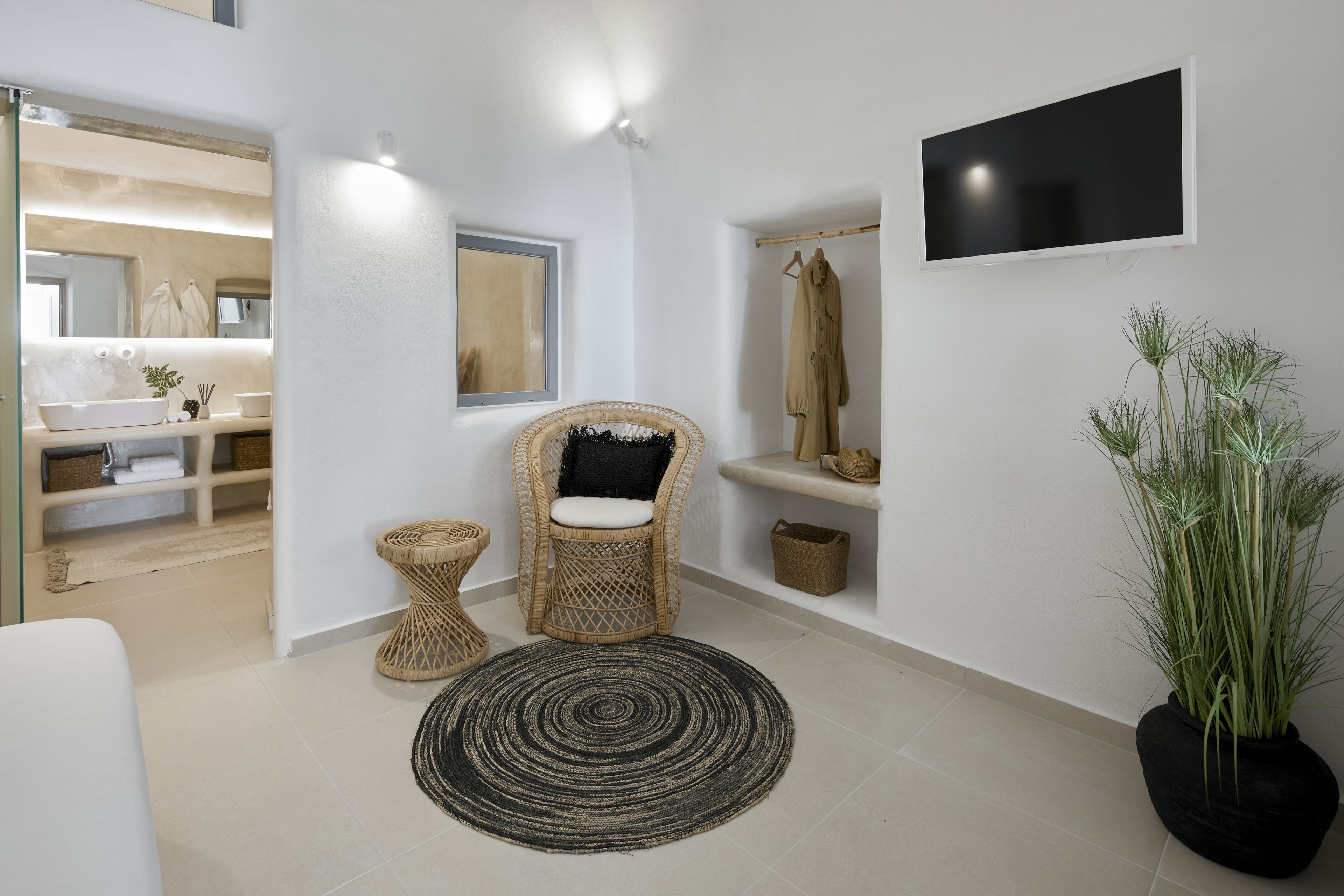 Un baño moderno con un espejo grande, un televisor de pantalla plana, una silla de mimbre, una planta en maceta y una alfombra circular.