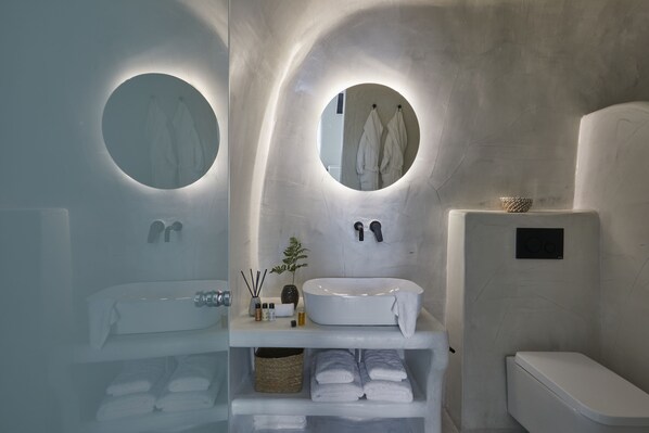 A modern bathroom with a white sink, two round mirrors, a toilet, and a shelf with towels and toiletries. - You And Me Suites (Santorini)