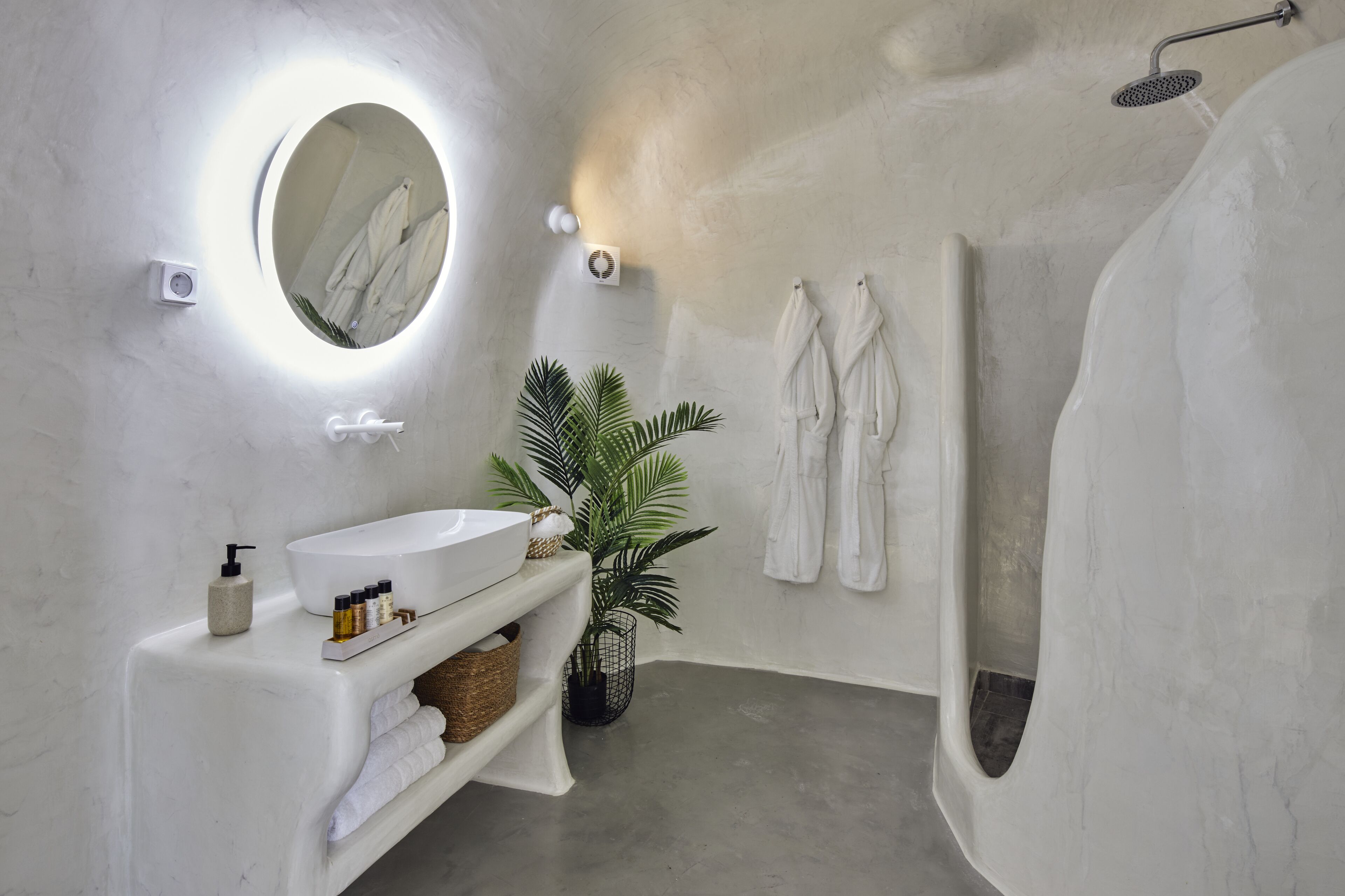 Un baño moderno con un lavamanos blanco, un espejo redondo con iluminación, zona de ducha y dos toallas blancas en la pared.