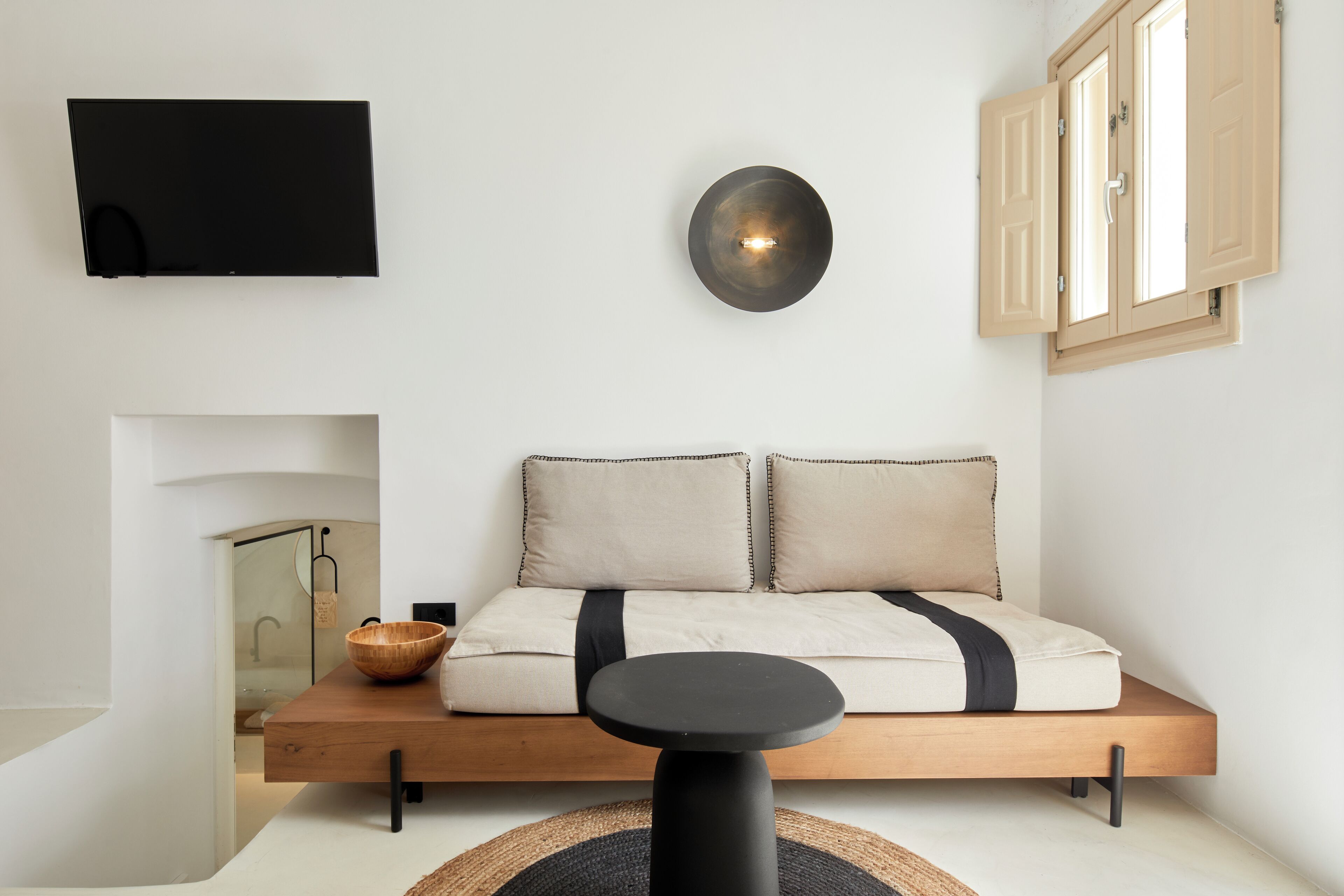 A modern room with a sofa bed, a mounted TV, a wall-mounted light, and a window with wooden shutters.