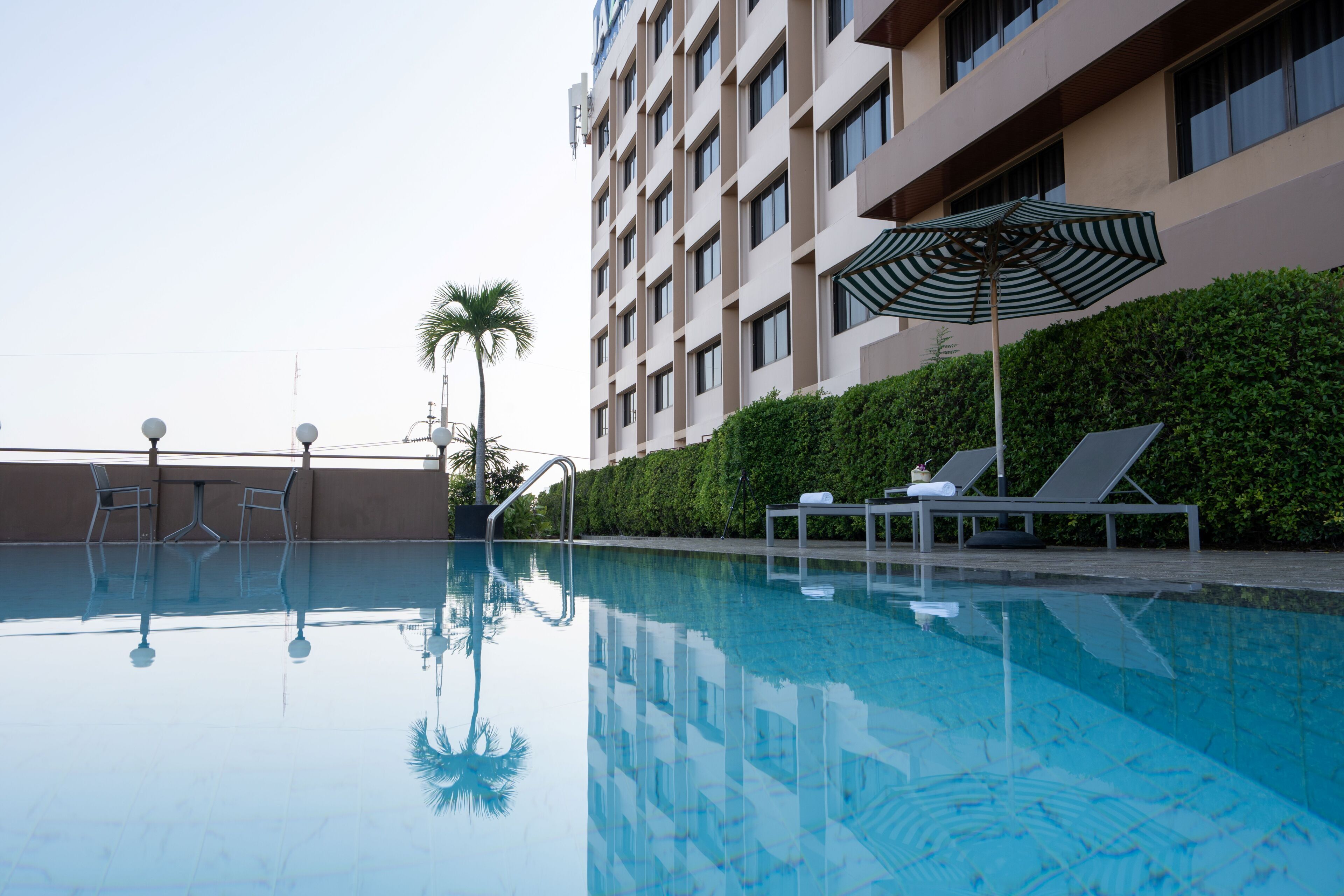 Una piscina con sillas de descanso y una sombrilla, rodeada por un edificio con varias ventanas y un seto.