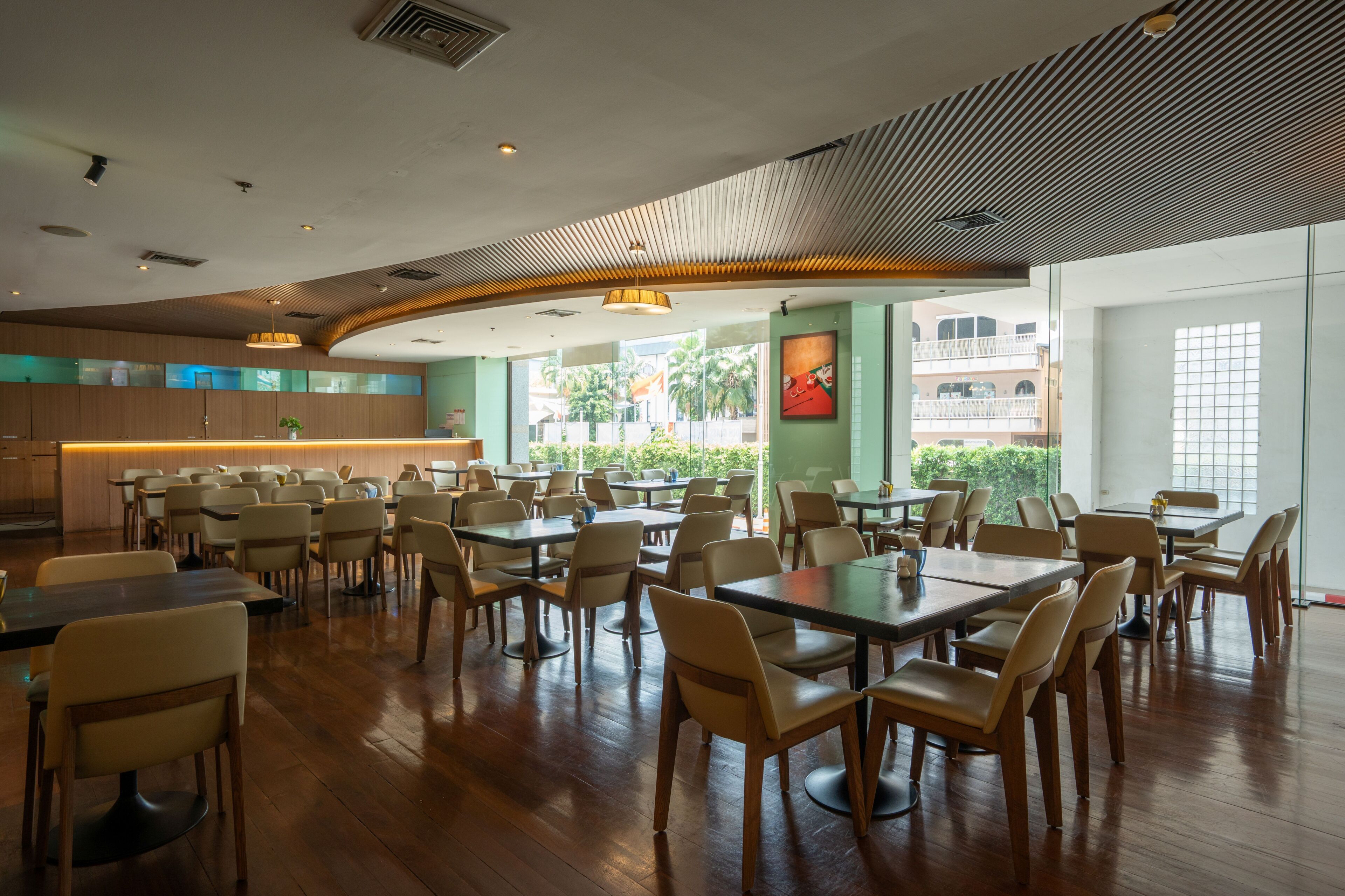 A spacious dining area with multiple tables and chairs, a kitchen in the background, and large windows providing natural light.