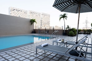 A rooftop pool with lounge chairs, a striped umbrella, and a view of a high-rise building.