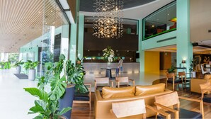 Un hall d’entrée spacieux au design moderne, comprenant un espace salon central avec des canapés en cuir et des fauteuils en bois, un grand lustre et une variété de plantes en pot.