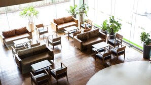 Un hall d’entrée spacieux, doté de plusieurs coins salon, comprenant des canapés et des fauteuils, disposés autour d’une table basse centrale. De nombreuses plantes en pot sont réparties dans tout l’espace.
