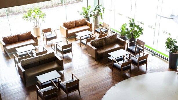 Un hall d’entrée spacieux, doté de plusieurs coins salon, comprenant des canapés et des fauteuils, disposés autour d’une table basse centrale. De nombreuses plantes en pot sont réparties dans tout l’espace.