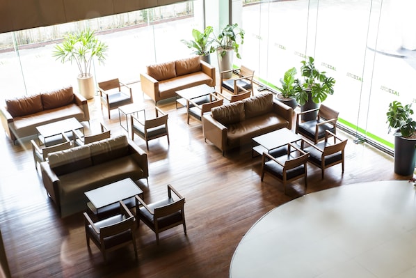 Un hall d’entrée spacieux, doté de plusieurs coins salon, comprenant des canapés et des fauteuils, disposés autour d’une table basse centrale. De nombreuses plantes en pot sont réparties dans tout l’espace.