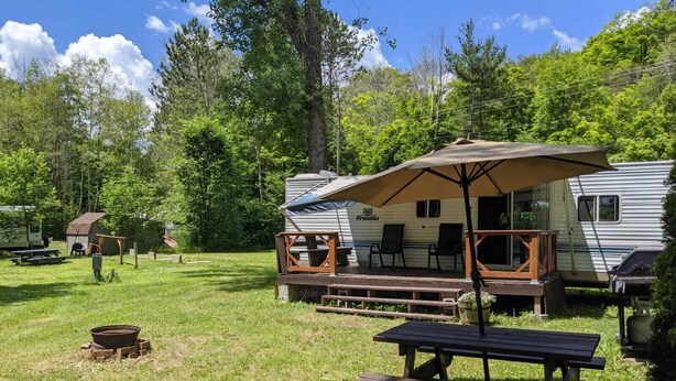 A campsite with a covered picnic table, a grill, and a camper with a porch. - Modern Rental Trailer at Bon Echo Family Campground (The Prowler) (Cloyne)