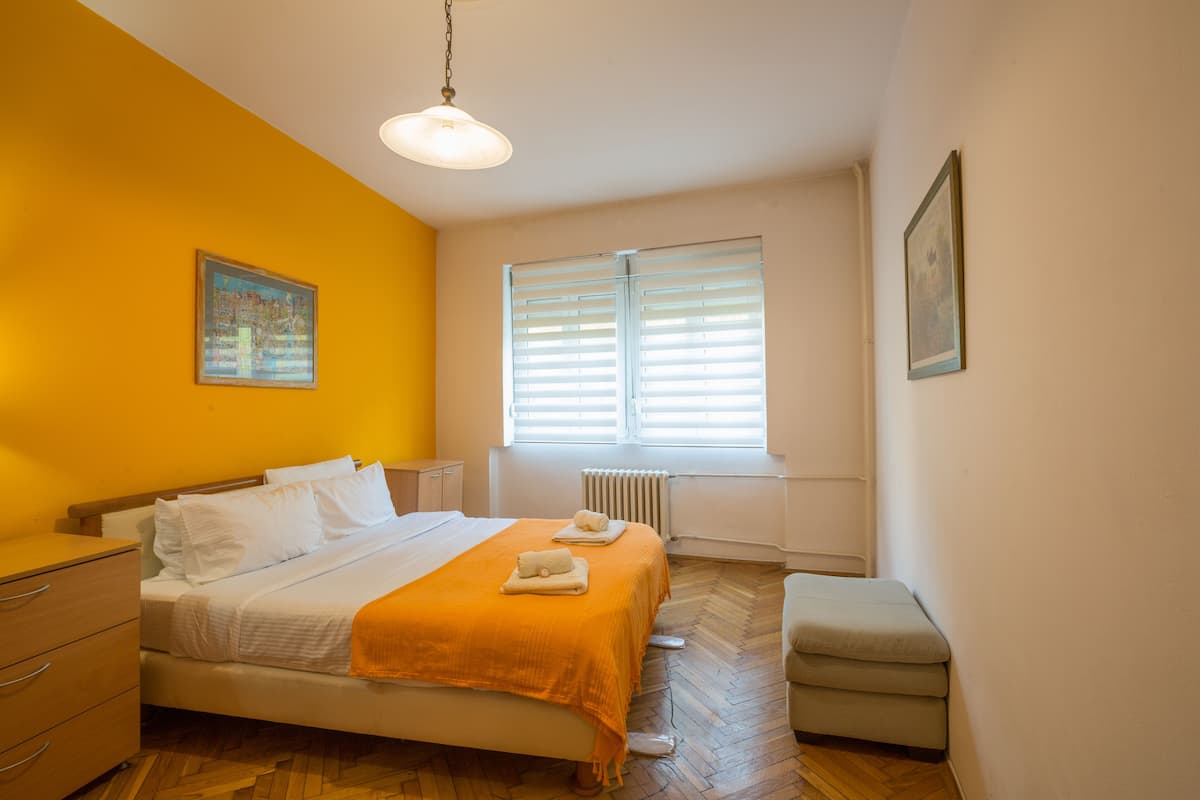 A bedroom with a bed, a nightstand, a dresser, a window with blinds, and a radiator.
