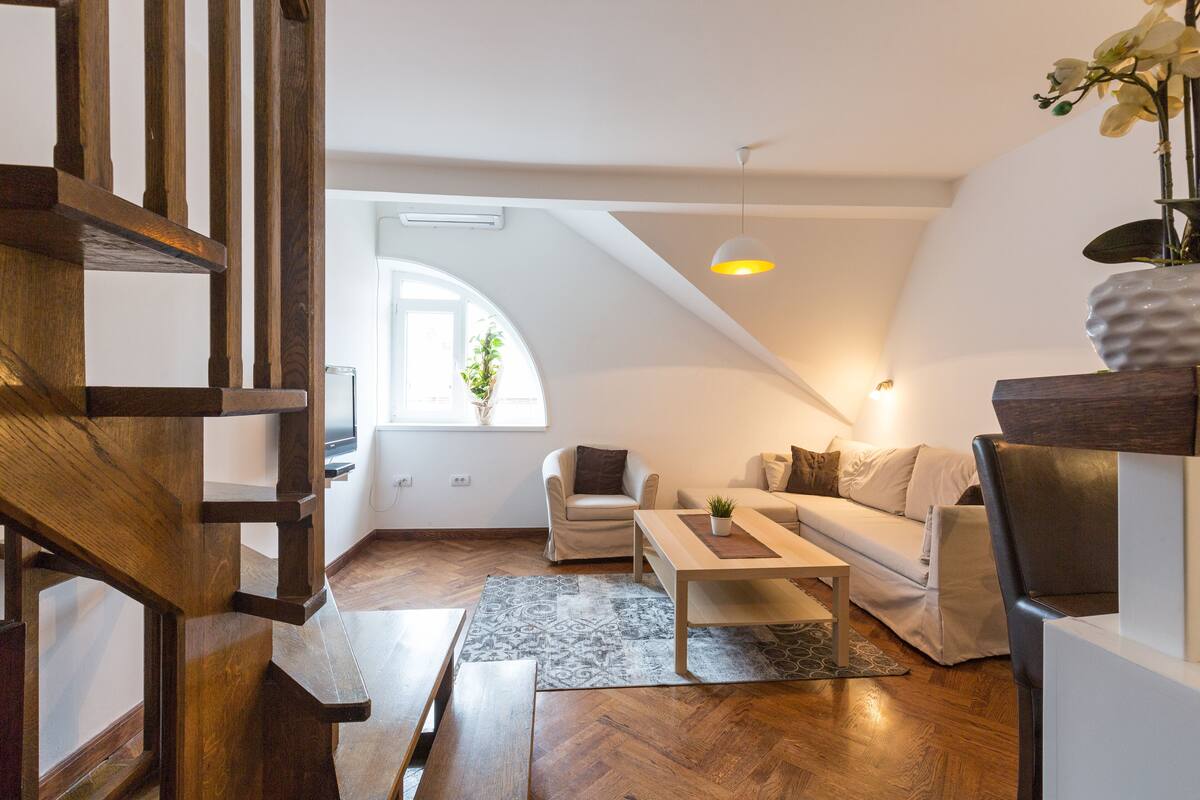 A modern loft apartment with a wooden staircase, a sofa set, a coffee table, and a hanging light fixture.