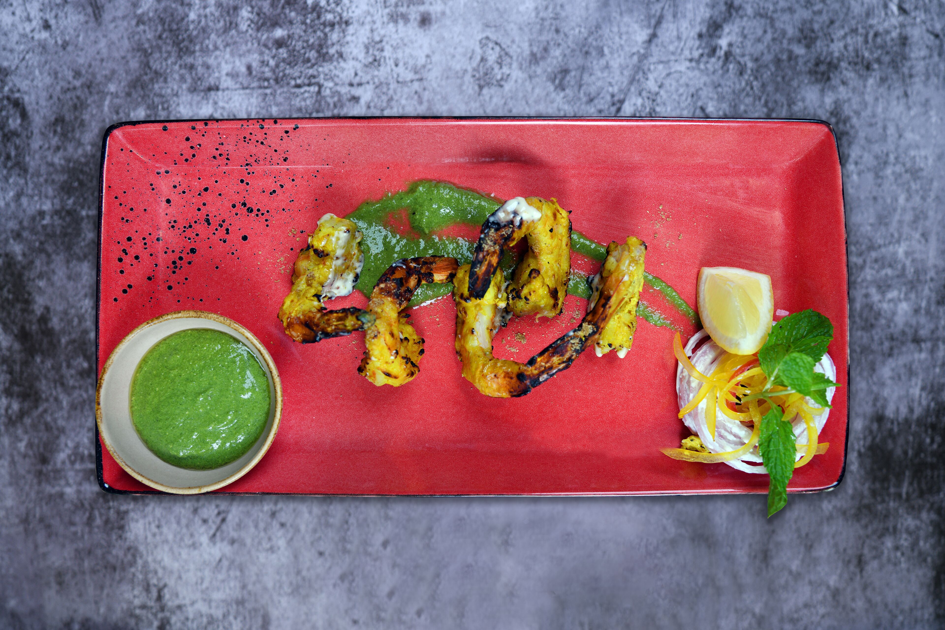 A rectangular plate with grilled vegetables, a bowl of green sauce, and a lemon wedge on a textured gray surface.