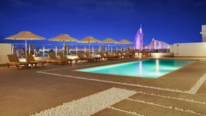 A rooftop pool area with lounge chairs and umbrellas, a clear view of a city skyline, and a distinctive building in the background. - The District Hotel (Dubai)