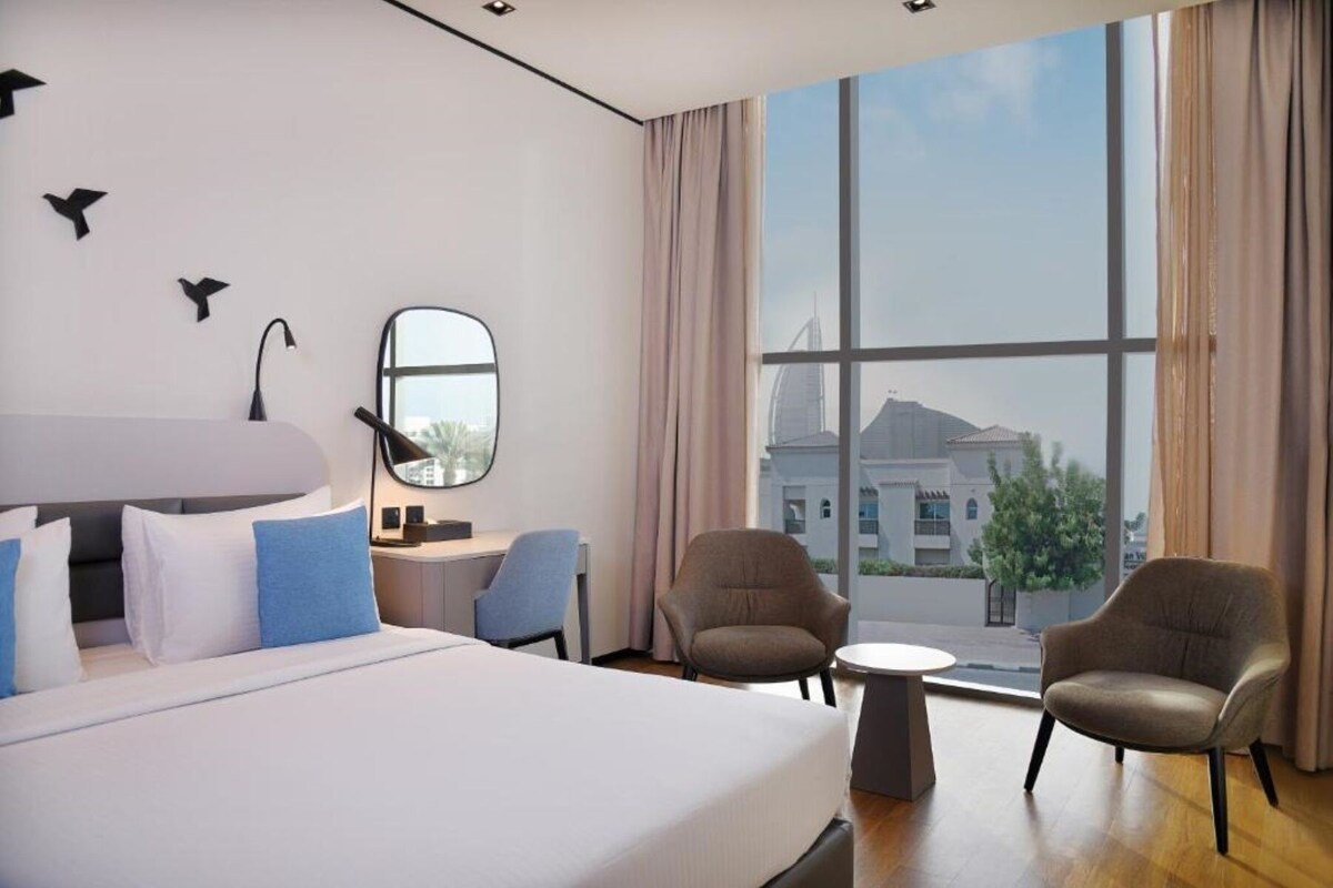 Ein modernes Hotelzimmer mit einem großen Bett, zwei Sesseln, einem Schreibtisch und Blick auf ein Gebäude durch große Fenster.