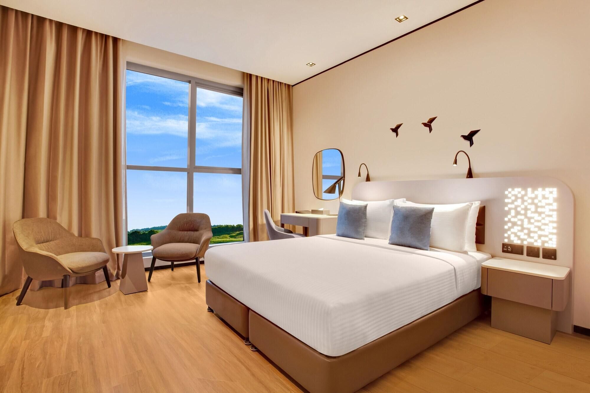 A modern hotel room with a large bed, two armchairs, a desk, and a view of the outdoors.