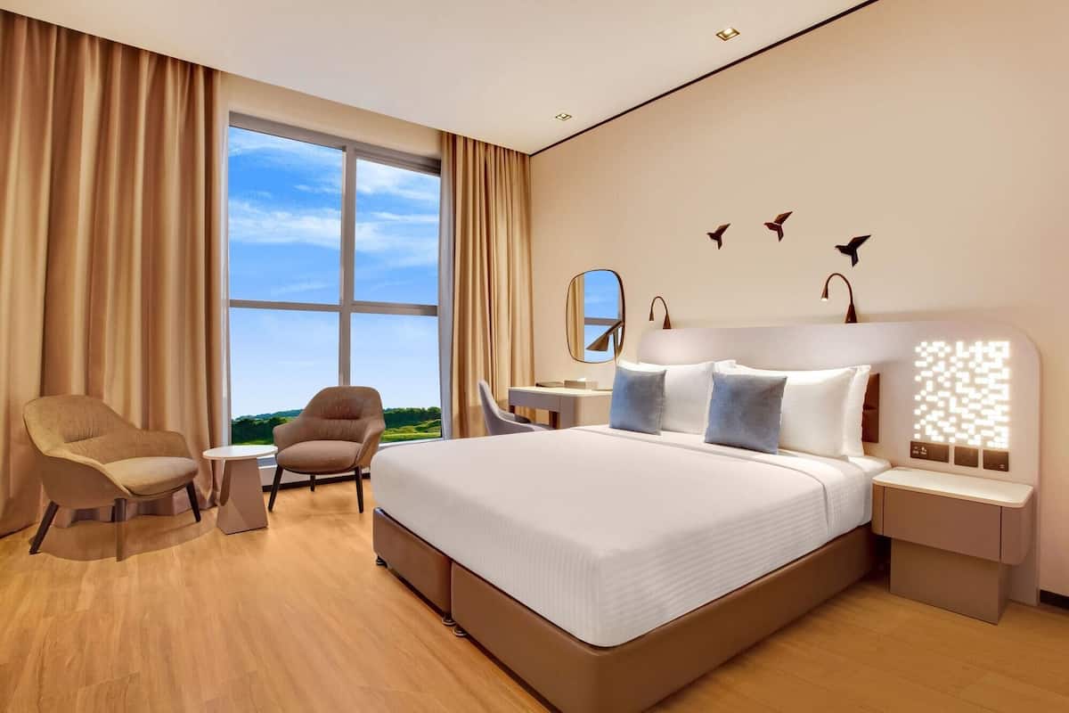 A modern hotel room with a large bed, two armchairs, a desk, and a view of the outdoors.