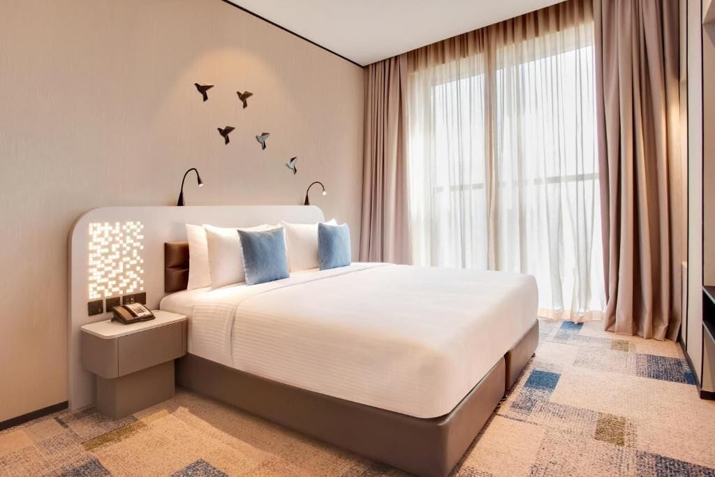 A hotel room with a large bed, a bedside table with a lamp, a wall-mounted phone, and a window with curtains.