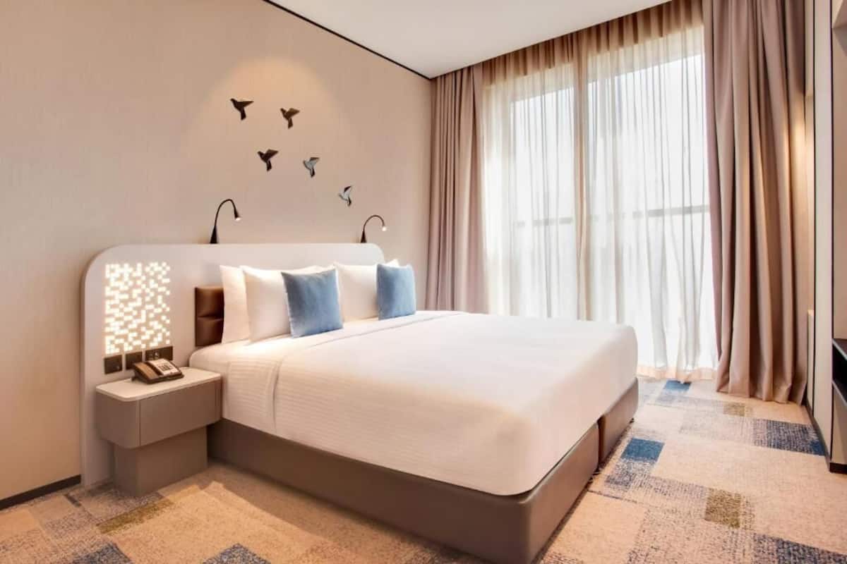 A hotel room with a large bed, a bedside table with a lamp, a wall-mounted phone, and a window with curtains.