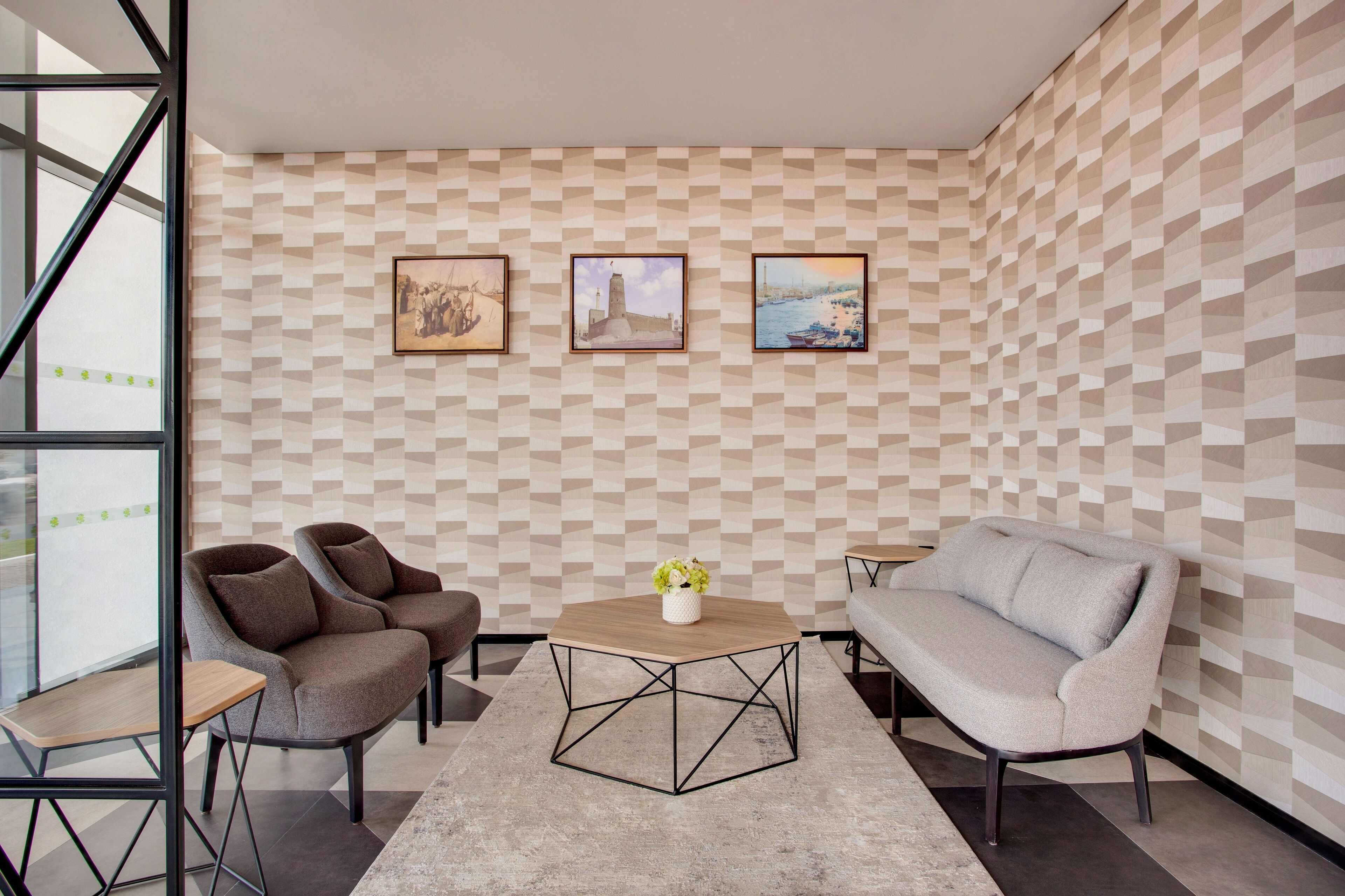 A modern room with a geometric patterned wall, a round wooden coffee table with a vase, two armchairs, and a sofa.