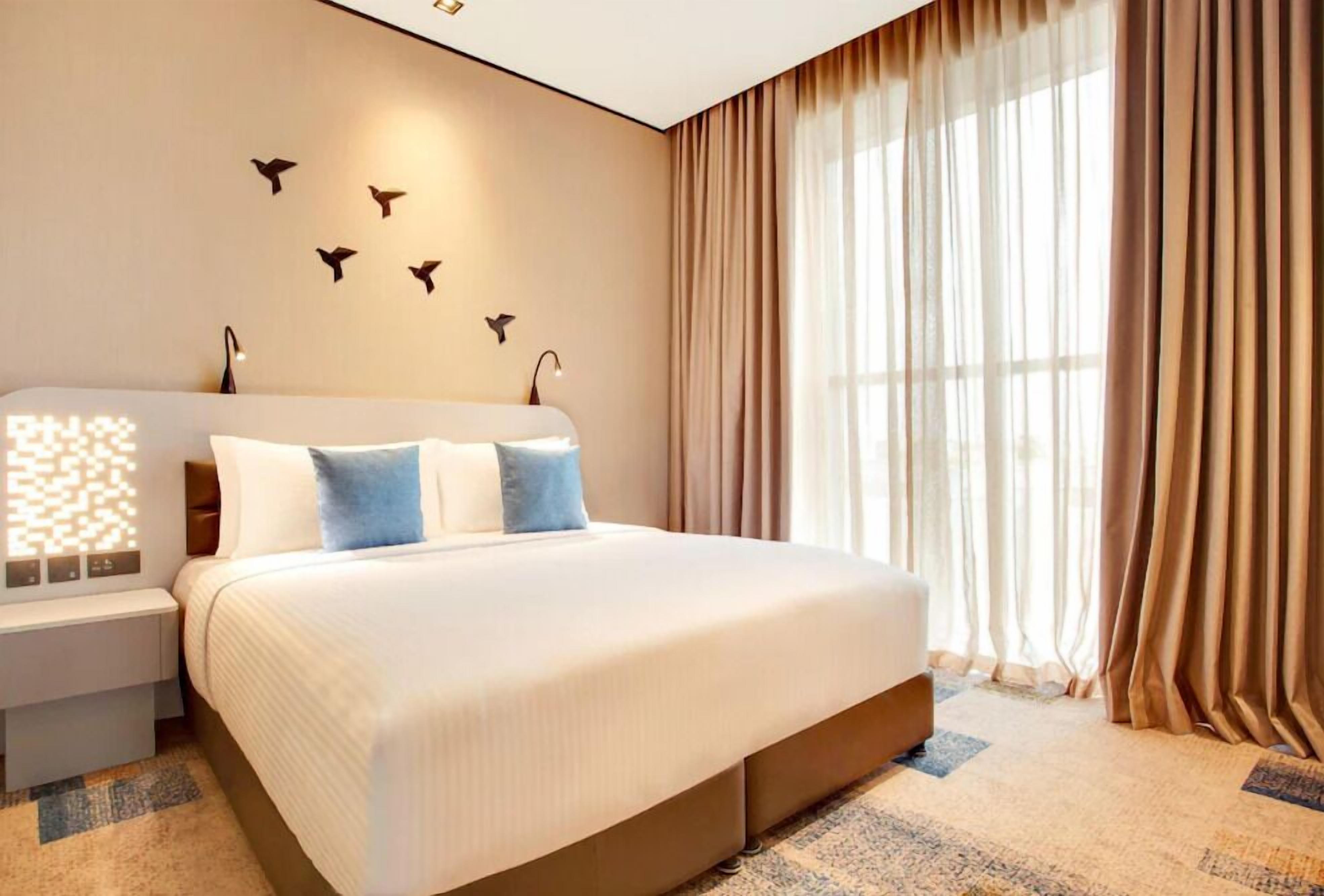 A modern hotel room with a large bed, a bedside table, a wall-mounted light, and a window with curtains.