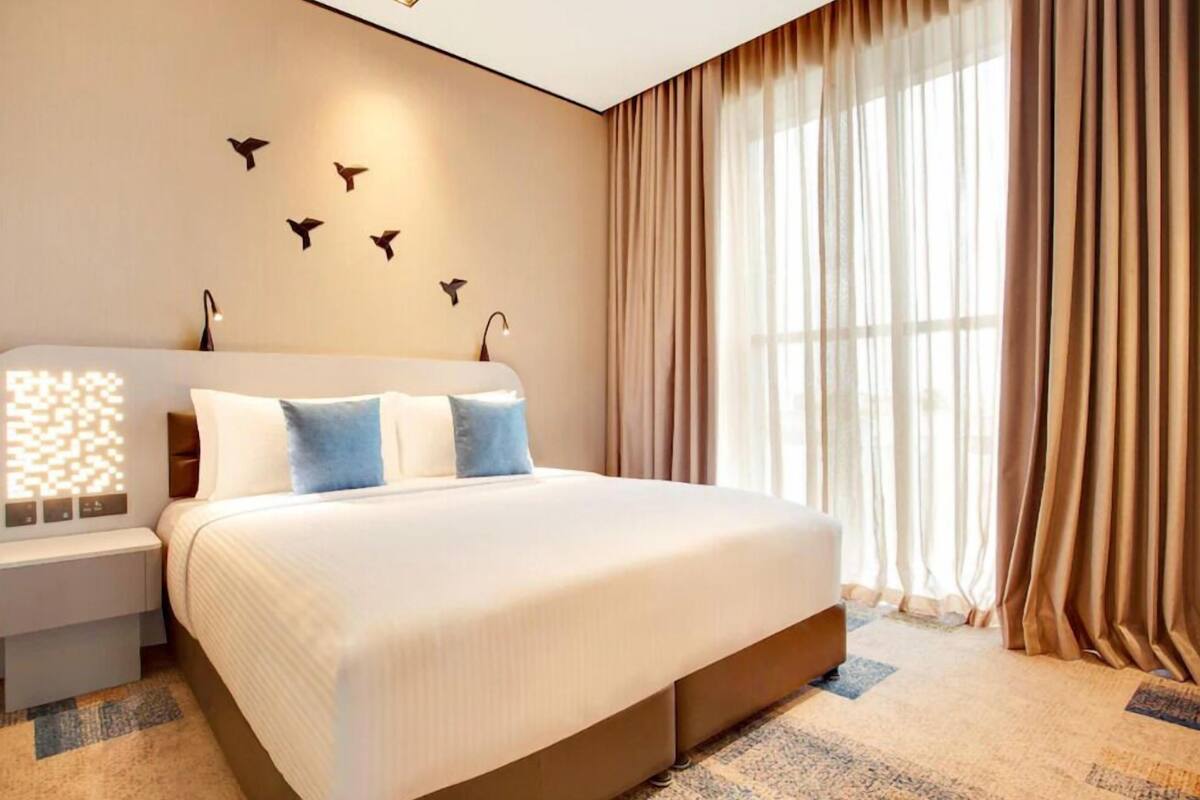 A modern hotel room with a large bed, a bedside table, a wall-mounted light, and a window with curtains.