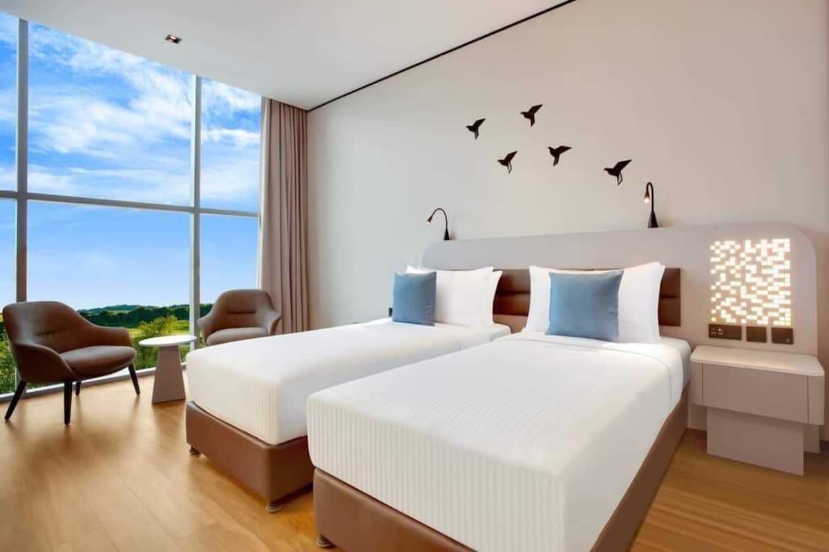 A modern hotel room with two beds, a large window with a view, a chair, and a wall-mounted light fixture.