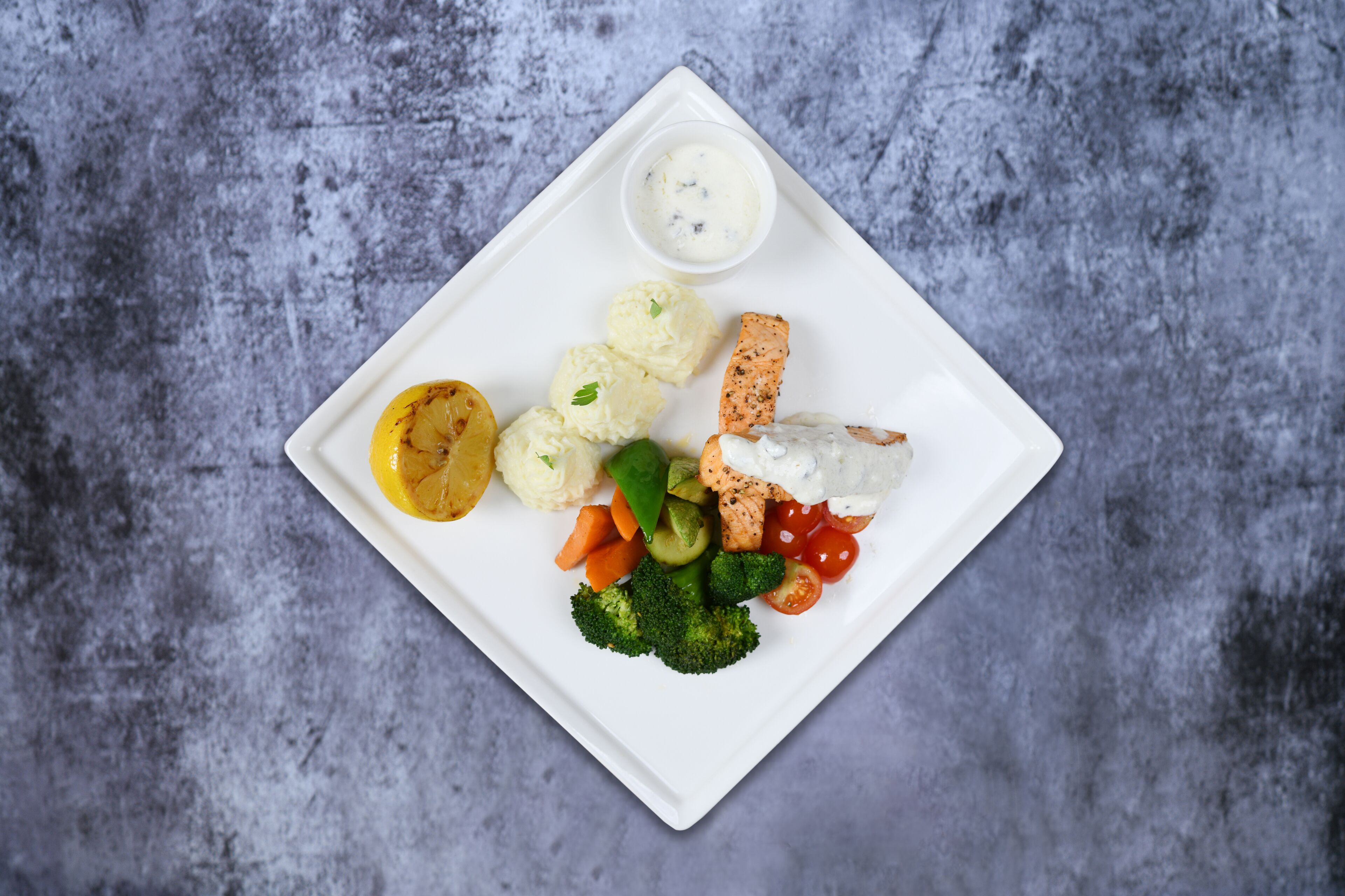 Une assiette carrée blanche contenant divers aliments, notamment du saumon, des brocolis, des tomates cerises et une portion de purée de pommes de terre.