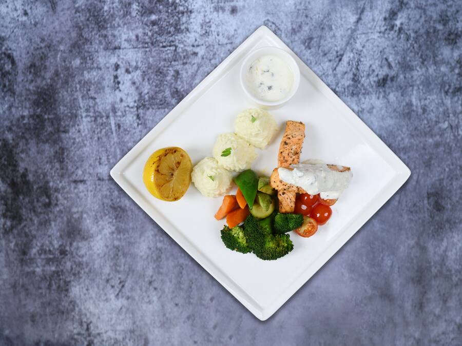 Une assiette carrée blanche contenant divers aliments, notamment du saumon, des brocolis, des tomates cerises et une portion de purée de pommes de terre.