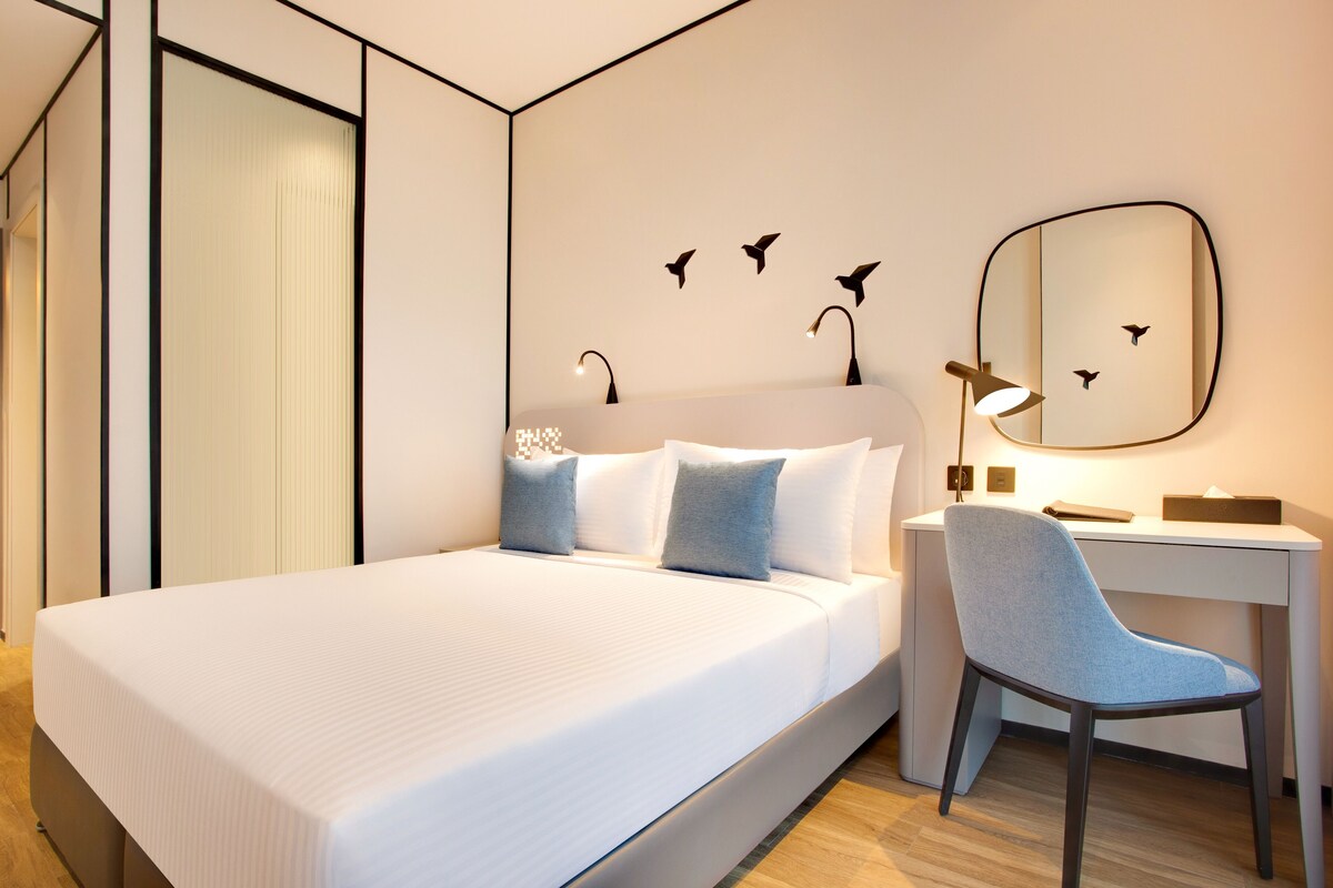 Ein modernes Hotelzimmer mit einem großen Bett, einem Schreibtisch und einem Stuhl. An der Wand befindet sich ein Spiegel und Kunstwerke mit Vogelmotiven.