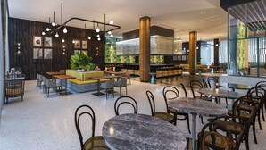 A modern indoor space with round tables and chairs, a central seating area with a sofa, and a large window providing natural light.
