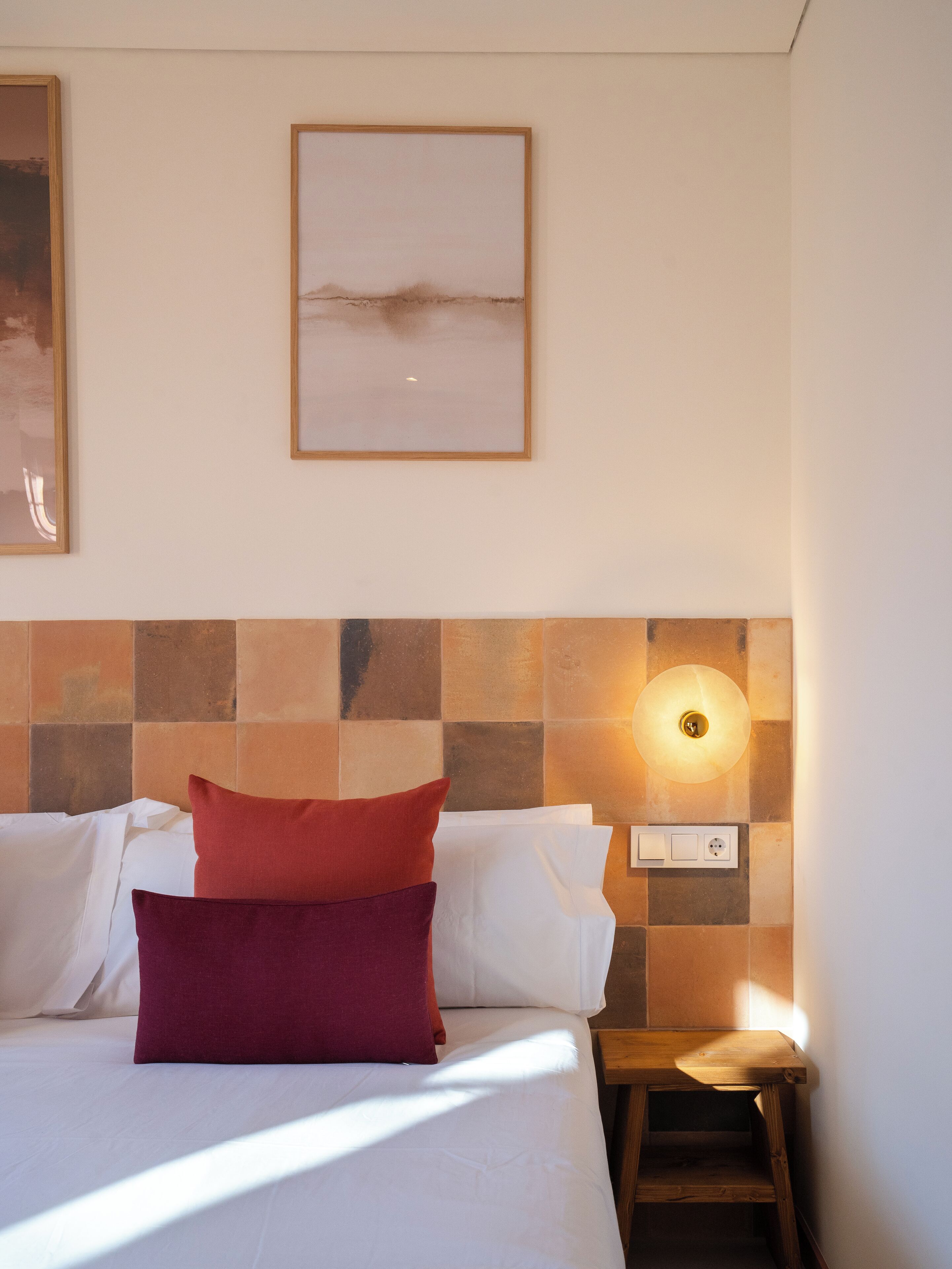 A neatly made bed with white and maroon pillows, a wall-mounted light fixture, and a small wooden nightstand.