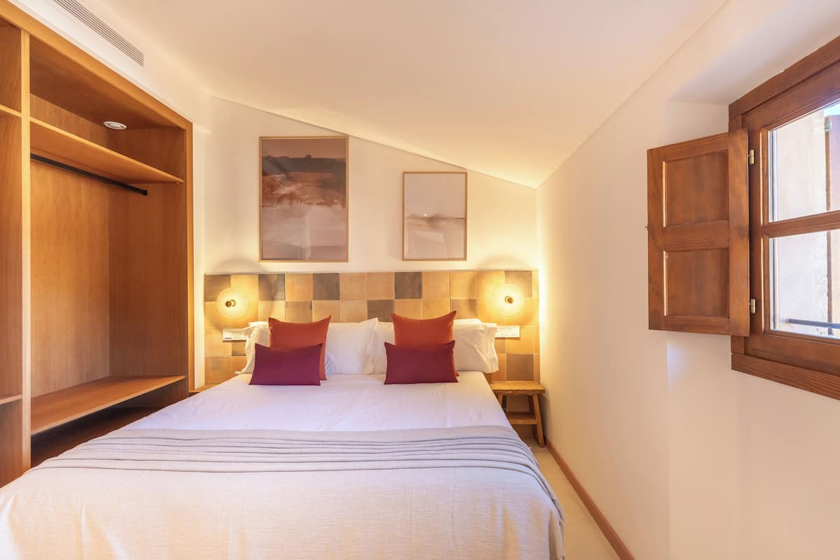 A hotel room with a large bed, wooden wardrobe, two wall-mounted lamps, and a window with wooden shutters.