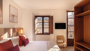 A bedroom with a bed, pillows, a chair, a TV, and a window with a view of buildings. - Marton Turisme de Interior (Valldemossa)