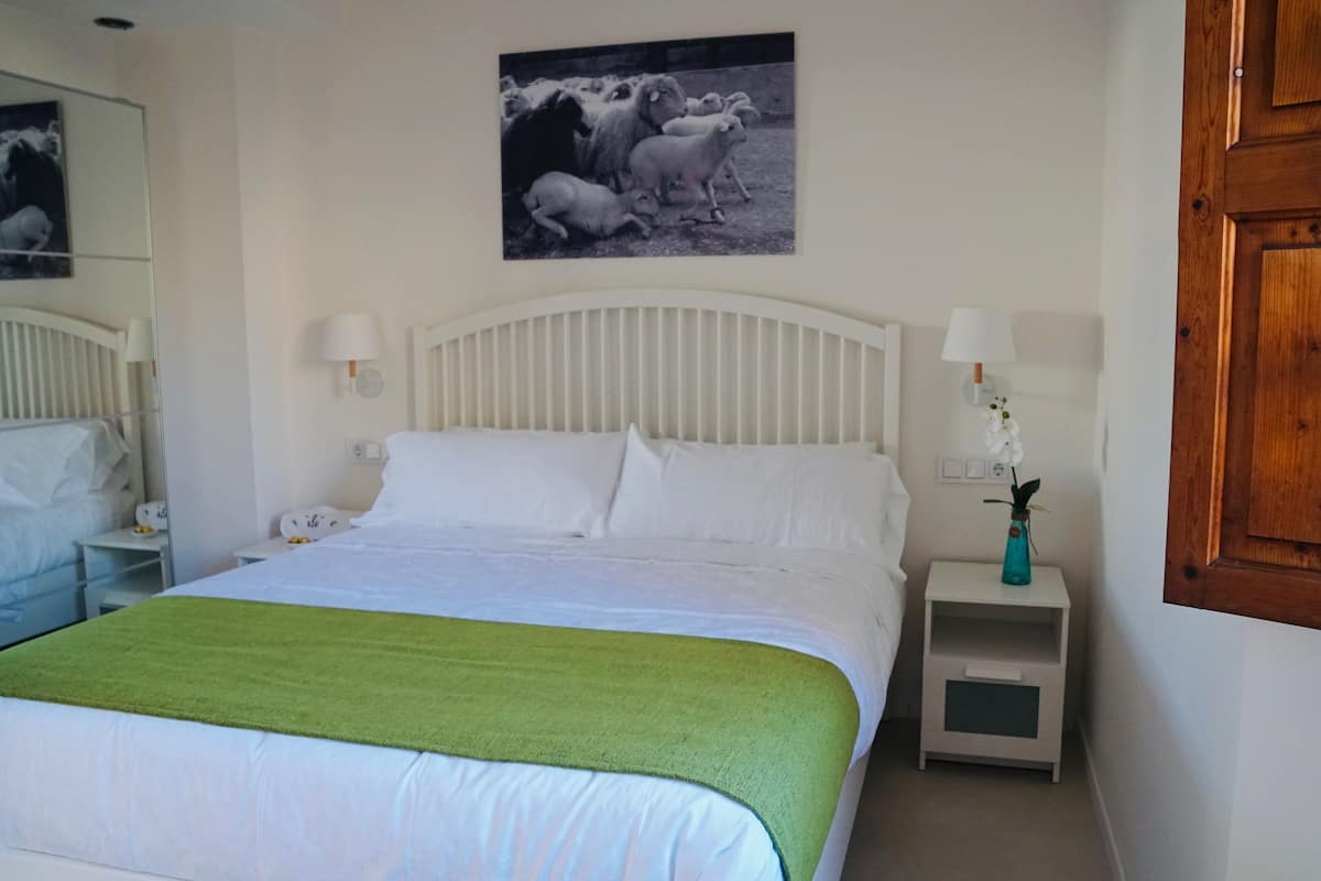 A neatly made bed with a green throw, a white headboard, two bedside lamps, a nightstand with a vase, and a wall-mounted picture of sheep.