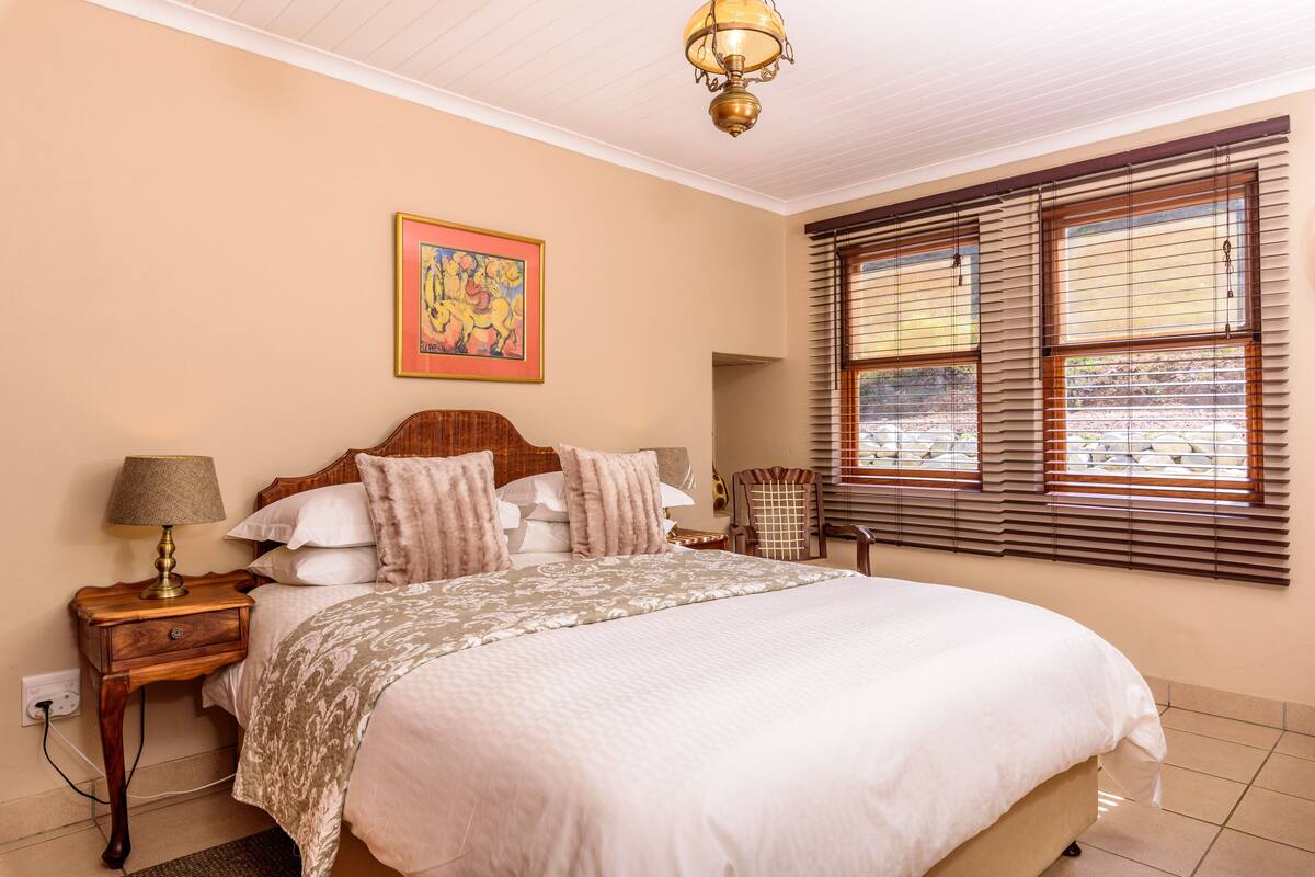 Een slaapkamer met een groot bed, een nachtkastje met lamp, een stoel en een raam met jaloezieën.