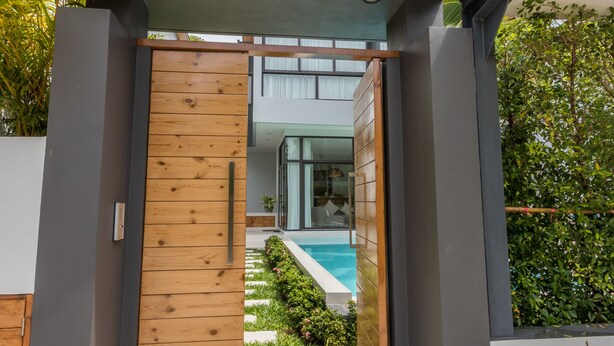 A modern house entrance with a wooden door, a concrete pathway, and a pool in the background. - 3 Bedroom Civetta Pool Villa (Rawai)