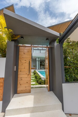 A modern house entrance with a wooden door, a concrete pathway, and a pool in the background.