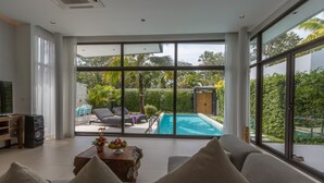 A modern living room with a large window overlooking a swimming pool and lush greenery. - 3 Bedroom Civetta Pool Villa (Rawai)