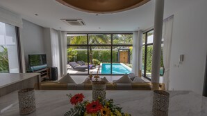 A modern living room with a marble table, a TV, and a view of a pool and garden. - 3 Bedroom Civetta Pool Villa (Rawai)