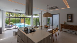 A modern kitchen with a central island, large windows, and a view of a swimming pool. - 3 Bedroom Civetta Pool Villa (Rawai)